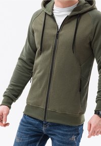 Felpa con cappuccio verde oliva con zip, realizzata in tessuto strutturato, dettagli a coste sulle maniche, coulisse regolabili e tasche laterali. Indossata sopra una maglietta bianca.