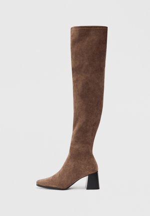 Botte hauteur genou en daim marron avec un talon bloc noir épais et un bout pointu sur un fond gris clair.