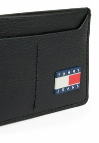 Portafoglio in pelle nera con superficie testurizzata, una tasca per carte e una placca rettangolare con logo con accenti rossi, bianchi e blu.