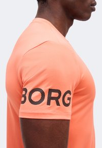 Πλάγια όψη ατόμου που φοράει ροδακινί κοντομάνικο πουκάμισο με μαύρο τελεία κείμενο "BORG" στο μανίκι.