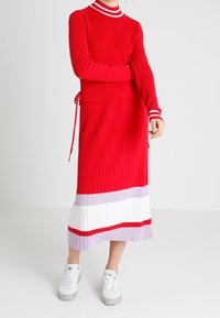 Roter gerippter Strickpullover mit einem weiß gestreiften Halsausschnitt, kombiniert mit einem rot, lila und weißen gestreiften Midirock, getragen mit weißen Sneakers.
