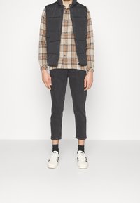 Chaleco acolchado negro sobre una camisa de cuadros beige y marrón, combinado con jeans oscuros y zapatillas blancas con rayas negras.
