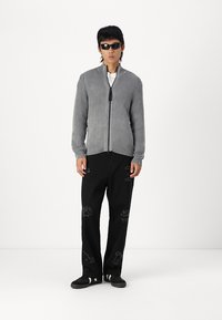 Suéter gris de canalé con cremallera combinado con pantalones gráficos negros que presentan patrones de garabatos y zapatillas negras sin cordones, todo sobre un fondo liso.