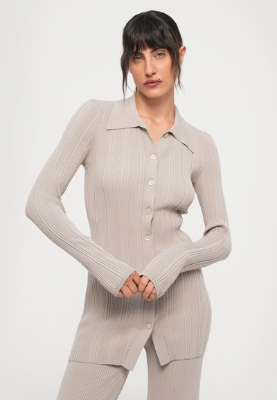 Woman with dark hair wearing beige ribbed long-sleeve buttoned cardigan and matching ribbed pants against a plain light background.
