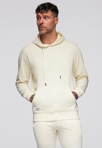 Homme aux cheveux bouclés et tatouages portant un sweat à capuche crème et un pantalon assorti, les mains dans la poche du sweat, regardant sur le côté.