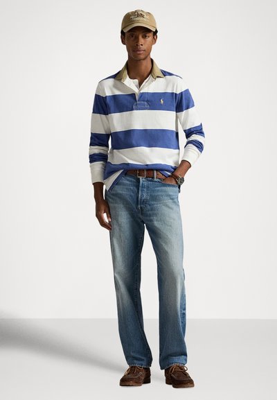 Muž nosiaci khaki čiapku, modro-bielo pruhované tričko s dlhým rukávom, svetlomodré rifle, hnedý opasok, hnedé topánky a náramkové hodinky, stojaci.