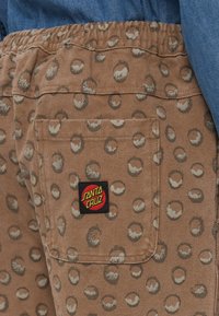 Brown fabric shorts featuring a pocket with a black “Santa Cruz” logo patch, and a circular pattern in light and dark shades.