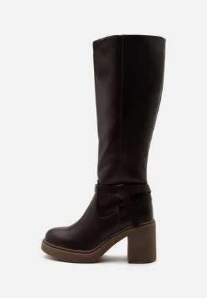 Hoher dunkelbrauner Lederstiefel mit Blockabsatz und dekorativem Riemen auf weißem Hintergrund.
