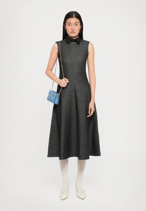 Sleeveless, knee-length dress in dark gray fabric with a collared neck. Model holds a small blue handbag with a chain strap. White tights and shoes.