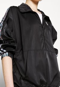 Černá lehká bunda s předním zipem, stojáčkovým límcem a elastickými manžetami. Má vzorovaný lem na rukávech a malé logo na hrudi.