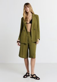 Donna che indossa un blazer testurizzato verde oliva e pantaloni corti fino al ginocchio, top bikini nero, sandali neri e bracciali robusti su un braccio.