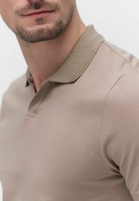 Beige polo skjorta med ett strukturerat tyg, en klassisk krage och placket med tre knappar. Den figurnära designen framhäver nacke och axelområde.