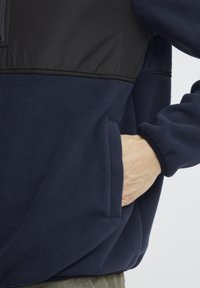 Giacca in pile blu scuro con una parte superiore in tessuto nero e liscio. Presenta una tasca frontale, polsini elastici e un design a vestibilità rilassata.