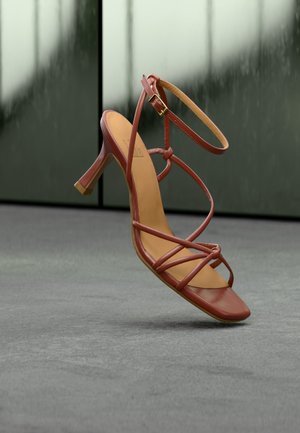 Καφέ ψηλοτάκουνο σανδάλι με λουριά και πόρπη στον αστράγαλο, που αιωρείται πάνω από γκρίζο τσιμεντένιο δάπεδο με θολό γυάλινο φόντο.