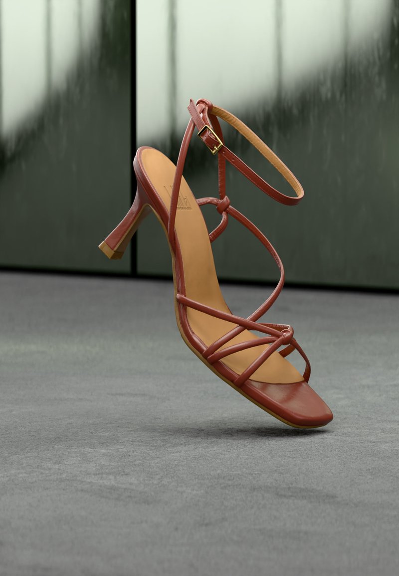 Καφέ ψηλοτάκουνο σανδάλι με λουριά και πόρπη στον αστράγαλο, που αιωρείται πάνω από γκρίζο τσιμεντένιο δάπεδο με θολό γυάλινο φόντο.