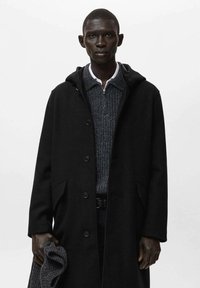 Abrigo negro con capucha, cierre de botones y bolsillos sobre un suéter gris de punto. Modelo sosteniendo una bufanda gris. Fondo blanco liso.