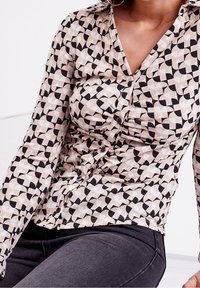 Mujer vestida con una blusa de botones arrugada y con un patrón de formas geométricas en beige, negro y blanco, combinada con pantalones gris oscuro.