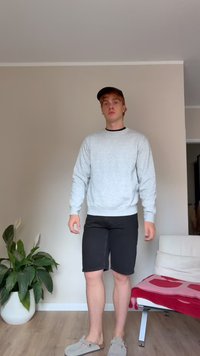 Felpa grigia con colletto nero, pantaloni corti neri e scarpe slip-on grigie. In piedi in una stanza con piante e mobili chiari.