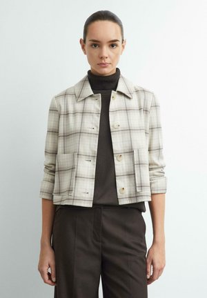 Frau, die eine hellbeige karierte Jacke über einem dunklen Rollkragenpullover und dunklen Hosen trägt, steht vor einem schlichten hellen Hintergrund.