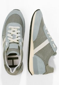 Paire de baskets en tissu gris clair et blanc avec des accents en daim, des lacets blancs et des semelles noires sur un fond blanc.