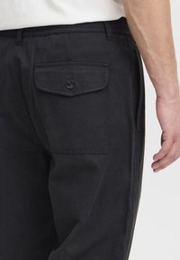 Pantalones de algodón negros con un bolsillo lateral con cierre de botón, costuras visibles y un diseño de ajuste relajado.