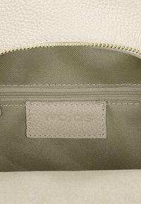 Beżowe wnętrze teksturowanej skórzanej torby z jasnobeżową podszewką z zielonkawej tkaniny, wyposażone w kieszeń na suwak oraz jasnobeżową etykietę z tłoczonym napisem "WOJAS".