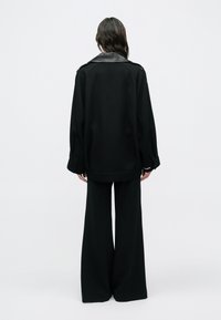 Čierna nadmerná bunda s koženým golierom, dlhými rukávmi a ozdobnými gombíkmi, doplnená o čierne široké nohavice, pohľad zozadu.