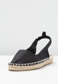 Espadrille en tissu noir avec un bout arrondi, semelle en jute tissée et bride arrière élastique. Présente des coutures blanches contrastées le long du bord.