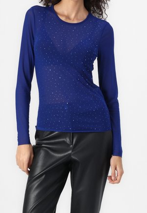 Mujer con una camiseta ajustada de manga larga azul real con pequeños cristales y pantalones de cuero sintético negro sobre un fondo liso.