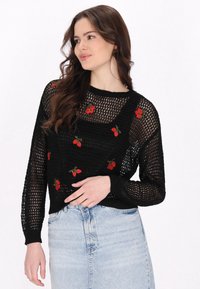 Schwarzer Strickpullover mit lockerem Maschenbild, verziert mit roten Erdbeer-Stickereien. Rundhals und lange Ärmel, kombiniert mit einem hellen Jeansrock.