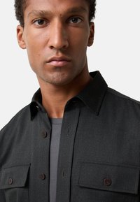 Black button-up shirt with two chest pockets, made of textured fabric. Features a classic collar and brown buttons, worn over a gray shirt.