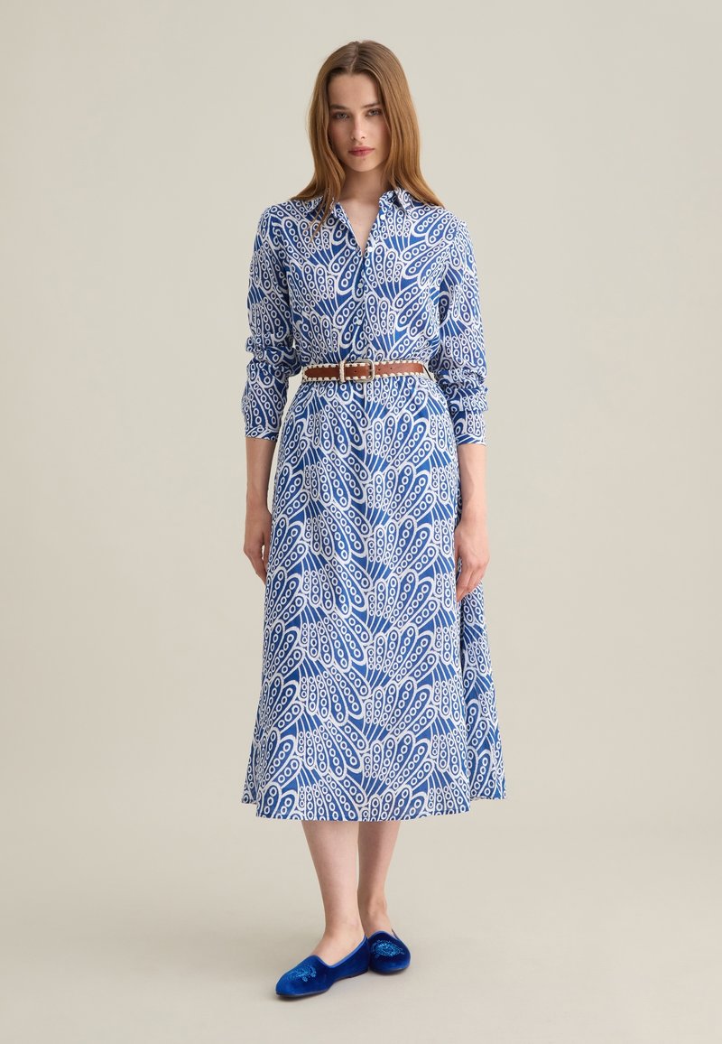 Lungo vestito blu e bianco con motivo, colletto, chiusura con bottoni, maniche a tre quarti e una cintura marrone. Indossato con scarpe basse blu.