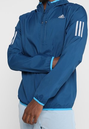 Leichte blaue Jacke mit Kapuze, ausgestattet mit silbernen drei Streifen an den Ärmeln und einer vorderen Reißverschlusstasche. Kontrastierende blaue Futter und Bündchen.