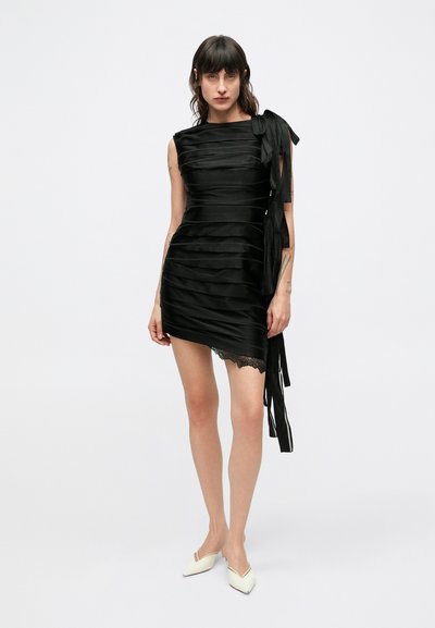 Čierne šaty s horizontálnymi saténovými skladmi, asymetrickým lemom, čipkovou lemovkou a mašľou na ramene. Nosia sa s bielymi topánkami so špicatou špičkou.