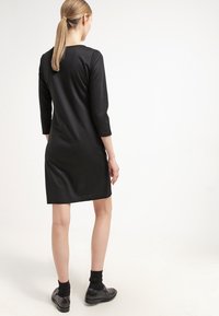 Svart klänning i mjukt tyg, med trekvartslånga ärmar och en enkel skärning. Bärs med svarta strumpor och skor, sedd bakifrån.