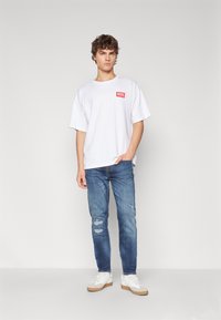 T-shirt blanc à manches courtes avec le logo rouge "DIESEL", associé à un jean bleu usé et des baskets blanches avec des semelles beige.