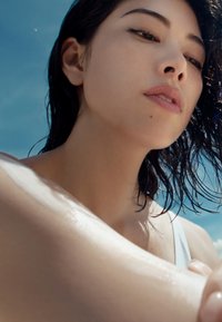 Gros plan sur une personne avec les cheveux et la peau mouillés, portant un maillot de bain de couleur claire, avec une épaule lisse et illuminée contre un ciel bleu.