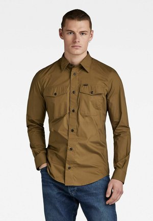 Chemise marron à manches longues en tissu lisse, dotée d'un devant à boutons, de deux poches poitrine et de boutons noirs. Associée à un jean bleu.