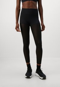 Persoon in zwarte high-waist leggings met doorzichtige panelen, zwarte sportschoenen en een zwart cropped sporttop op een effen achtergrond.