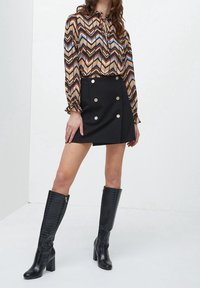 Zkrácená hnědo-modrá halenka se vzorem cikcak, černá minisukně se zlatými knoflíky a černé kozačky až ke kolenům s krokodýlím efektem.