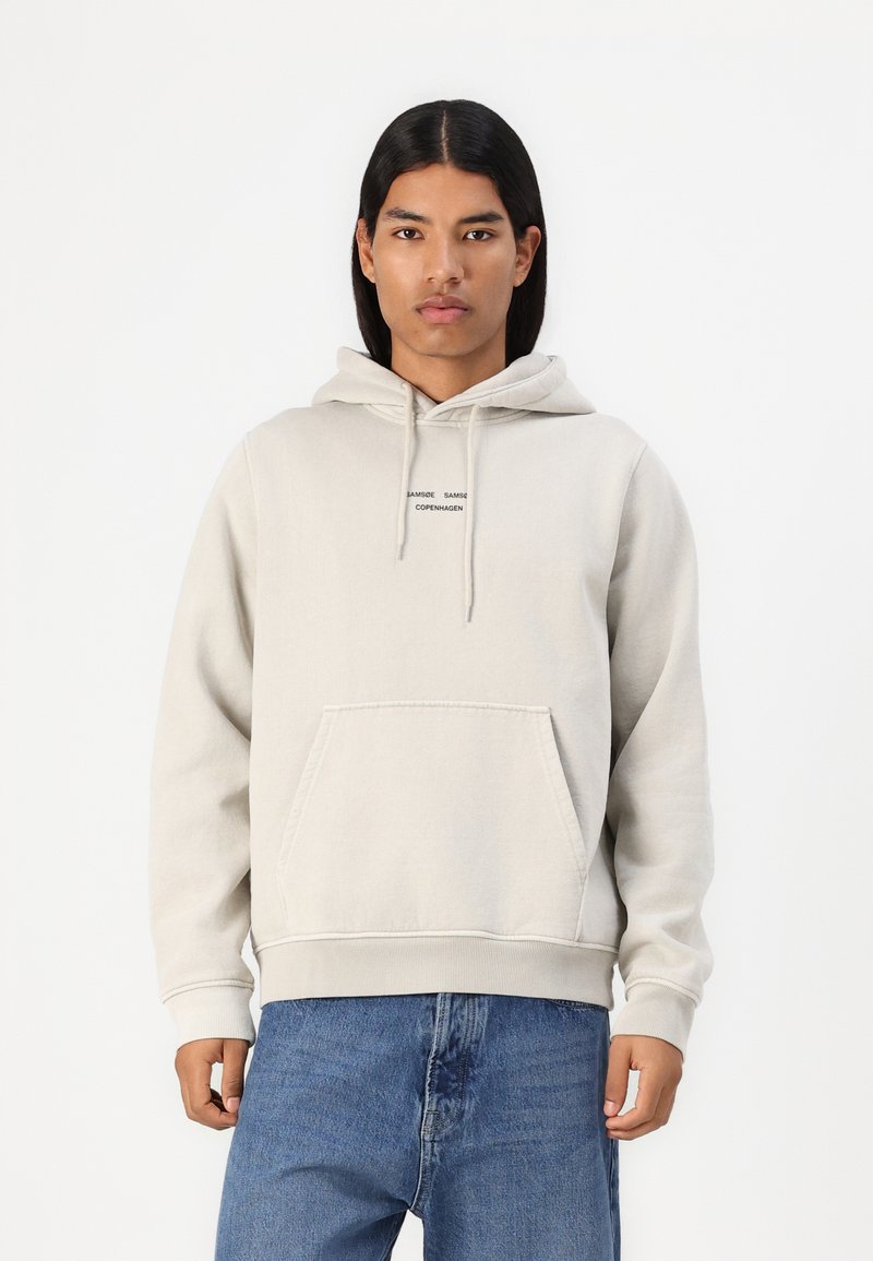 Lys grå hoodie med snor i hætten, forlommer og ribbede manchetter. Har en lille sort logo tekst på brystet. Bæres over denimjeans.