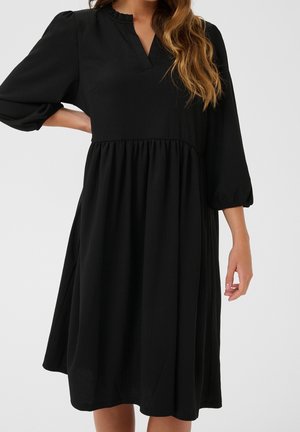 Mujer que lleva un vestido negro suelto hasta la rodilla con mangas globo hasta el codo y un pequeño detalle de cuello en V.