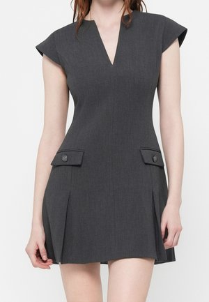 Frau trägt ein kurzes, dunkelgraues Kleid mit Kappärmeln, V-Ausschnitt und Klapptaschen mit Knöpfen an den Hüften.