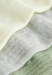 Három réteg texturált kötött anyag halvány sárga, világosszürke és lágy zöld színekben, mindegyiken kerek, kiemelkedő pöttyök tapintható részletekkel.