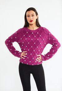 Fuchsia strikket sweater med et diamantmønster, dekoreret med hvide perler langs sømmene og ærmerne. Lange ærmer, runde halsudskæring.