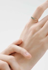 Anillo de oro con múltiples pequeñas piedras preciosas azul-verdosas y acentos claros, llevado en una mano con piel suave. Diseño sencillo con banda delgada.