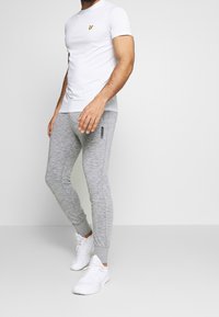 Hvid kortærmet t-shirt med et lille gult logo på brystet, parret med lysegrå tætsiddende sweatpants og hvide sportsko.