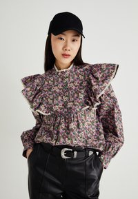 Bluse med blomsterprint og store volangermer, med høy krage og lacedetaljer. Båret med svarte skinnbukser og en svart caps.
