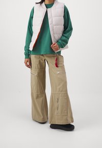 Puffige weiße Weste über einem grünen Langarmshirt, kombiniert mit beigen Cargo-Hosen mit mehreren Reißverschlüssen und einem roten Etikett-Detail. Schwarze Schuhe.