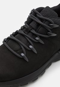 Czarne buty trekkingowe z cholewką z zamszu i siateczki, gumową podeszwą oraz teksturowanymi czarnymi sznurowadłami zabezpieczonymi plastikowymi blokadami.